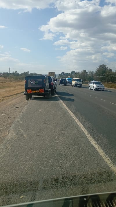 A sunny day scene features a highway with multiple vehicles, including a black tuk-tuk-like trailer with license plate Z6 0307 parked on the left side. Cars and vans fill the lanes going the same direction. Blue skies with scattered clouds dominate the background. A dry grassy area and some trees border the highway. The image is taken from inside a vehicle, looking out across the road.