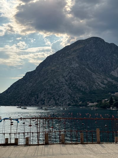 The image depicts a waterfront scene in Donji Orahovac, Montenegro, dominated by a large, rocky mountain that slopes steeply into a dark body of water. The sky is partly cloudy, with patches of sunlight breaking through dramatic grey clouds. In the foreground, a concrete structure with exposed rebar forms a fence-like barrier, with several vertical wooden posts and horizontal rebar reinforcing it. Attached to the rebar are numerous small, red and orange buoys, strung together by thin lines. The water itself is choppy and reflects the sky's light. A few boats, including sailboats and motorboats, are visible on the water further out, and the shoreline on the far side of the bay is lined with trees and scattered white buildings, suggesting a small settlement. The overall impression is of a rugged, natural landscape contrasted with an ongoing construction or development project at the water's edge. There are no people visible in the frame.