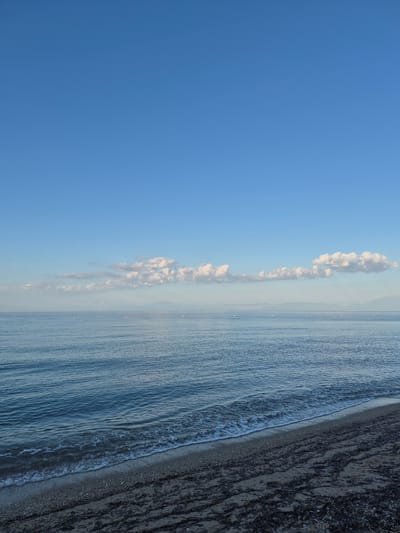 This is a vertical, eye-level shot of a serene seascape at Νέα Καλλικράτεια, Greece. The image is dominated by a vast expanse of clear, deep blue sky that occupies the top two-thirds of the frame. A thin band of light, hazy clouds stretches across the horizon line, where the sky meets the calm, blue sea. The water exhibits gentle ripples, creating a textured surface. Along the bottom third of the image, a dark, pebble-strewn beach is visible. Scattered dark seaweed lies on the sand, and the edge of the sea is marked by a gentle, foamy wave lapping the shore. In the distance, faint outlines of mountains are visible against the horizon. The scene is tranquil and devoid of any human presence or activity.