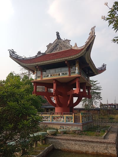 Captured in the city of Lumbini Sanskritik, Nepal, this outdoor shot features a red pagoda-style structure with an ornate, tiered roof. The roof is adorned with traditional dragon and phoenix motifs. The building has a second-story balcony and is elevated on a multi-tiered base with decorative pillars and supporting arches. Below, a small, landscaped courtyard surrounds a murky pond or pool. Vegetation, including trees and bushes, is visible in the foreground and surrounding area. In the background, construction structures can be seen, indicating the presence of ongoing development. The scene is bathed in soft, natural light, suggesting either early morning or late afternoon.
