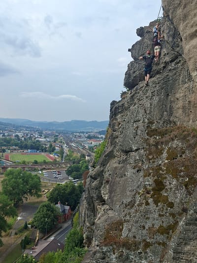 The image shows three people rock climbing on a sheer cliff face with a scenic overlook of Děčín, Czechia. The climbers, equipped with helmets and harnesses, are ascending a via ferrata, a protected climbing route that uses steel cables, rungs, and ladders. The person in the foreground, wearing a black shirt and shorts, is facing away from the camera, rappelling or descending down the rock face. Behind them, two individuals, one in a white and blue top and the other in a darker top and beige shorts, are climbing upward. The rock face is rough and weathered, with patches of moss and vegetation. In the background, the city of Děčín sprawls across the landscape, featuring a stadium, railway tracks, buildings, and a distant mountain range under a cloudy sky. A red brick building with a domed roof is visible closer to the foreground, nestled amongst trees. Cars are parked in a lot near the stadium, and a bridge with arches spans a road.