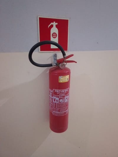 A red fire extinguisher is mounted on a light-colored wall. Above the extinguisher is a square sign with a white outline, a red background, and a white silhouette of a fire extinguisher with "PÓ ABC" written in a white circle on it. The fire extinguisher itself is red and has a black hose and a silver nozzle. There is a white label on the extinguisher with some text and graphics. The wall appears to be indoors, possibly in a hallway or public space. There are no people visible in the image. The location is Cáceres, Brazil.