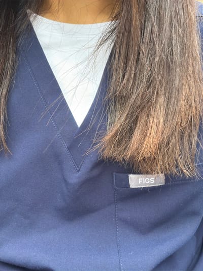 A close-up, angled shot captures the upper body of a person, primarily focusing on their hair and clothing. The dominant subject is a mass of long, dark brown hair with visible lighter brown or reddish highlights, appearing somewhat textured and flowing. Part of the person's skin, a light to medium brown tone, is visible on the left side of the frame, suggesting the neck or ear area.
The person is wearing a navy blue garment, likely a medical scrub top, characterized by a visible V-neck and clear stitching details along its edges. A plain white fabric, possibly an undershirt, is seen beneath the V-neck of the blue top.
A key detail is a rectangular brand tag sewn onto the blue garment on the upper right side. The tag is gray with the word "FIGS" printed vertically in white, capitalized letters. The fabric of the blue garment exhibits a fine, diagonal weave pattern. The lighting is bright and even, emphasizing the textures of the hair and clothing. The image is static, depicting the person wearing the uniform, with no specific activity taking place. The setting appears to be indoors or in a controlled environment, with no discernible external location cues or time of day.