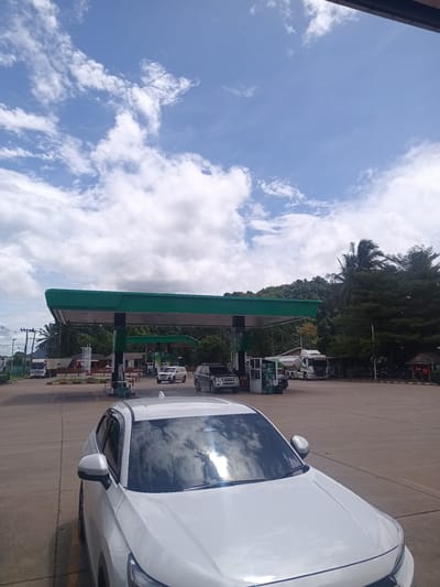 The image captures a sunny day at a PTT gas station in Plai Phraya, Thailand. A white car dominates the foreground, reflecting the cloudy sky on its windshield. The gas station features a green canopy covering the pumps, where multiple vehicles, including trucks and SUVs, are visible. Lush green trees line the background, adding to the outdoor setting. The sky is partially covered with white clouds against a blue backdrop.