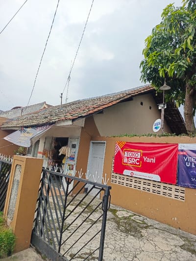 A tan-colored building with a tiled roof features a small shop at the front. A red "TOKO SRC Yani" banner hangs on the wall beside a white door. An Aqua sign is mounted higher up. A black iron gate stands in front of a concrete walkway, with green grass visible at the edges. A large tree with green leaves is on the right. Overhead wires stretch across the cloudy sky.