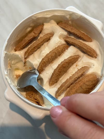 An overhead view within a domestic setting in Kalyvia Thorikou Municipal Unit, Greece, depicts a white rectangular container partially filled with a light-colored, creamy dessert substance, possibly ice cream or yogurt. Approximately ten elongated, ridged, light brown biscuit-like pieces are arranged mostly parallel to each other on the surface of the dessert. A silver-colored metal spoon is positioned in the left side of the container, partially submerged in the dessert, with a portion of the dessert displaced, revealing a lighter, possibly yellow, layer underneath. A human hand, exhibiting manicured fingernails with light pink polish, grasps the handle of the spoon from the lower right quadrant of the image, indicating an active consumption of the dessert. The background consists of a light-colored, possibly white or off-white, horizontal surface.