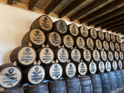 The image shows a large collection of wooden barrels stacked indoors, likely in a distillery or storage facility. The barrels are arranged in several tiers, with the front-most barrels clearly visible. Each barrel has a circular marking on its end, featuring the text "SINCE 1590 STABKA ZAPOLSKAYA BELARUS" and a stylized emblem. The barrels vary in color, from light to dark wood, and have visible metal bands around them. The interior environment appears rustic, with a wooden beam ceiling and a white wall. The lighting suggests an indoor setting, possibly with natural light. No people are present in the image. The location is identified as Litvensky Sel'skiy Sovet, Belarus, which aligns with the "BELARUS" inscription on the barrels.