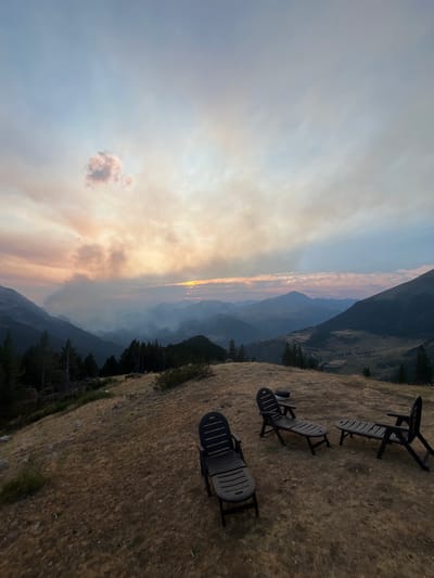 The image depicts a serene mountain landscape at dusk. In the foreground, three dark-colored lounge chairs are arranged on dry, golden grass. Beyond the chairs, a dense forest of dark green pine trees lines the crest of the hill. The middle ground features a valley filled with layers of hazy, blue-tinged mountains, with a hint of smoke or fog obscuring the lower slopes. The sky is a gradient of soft blues and pinks, with streaks of clouds catching the fading sunlight, suggesting either sunset or sunrise. A single, small pink cloud is visible in the upper left quadrant of the sky. The overall atmosphere is peaceful and remote, characteristic of a mountain viewpoint in an area like San Juan de Plan, Spain, known for its natural beauty and mountainous terrain.