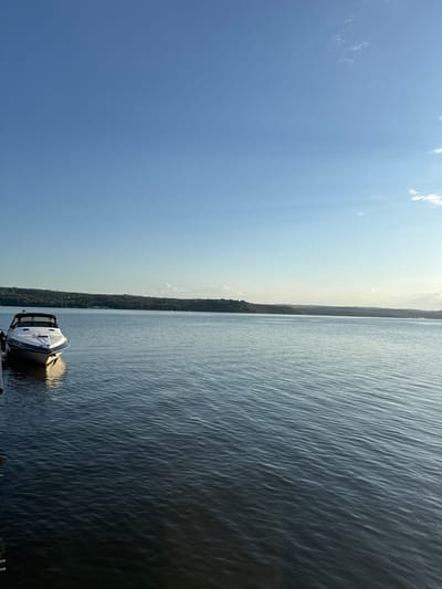 A white and blue motorboat is docked on the left side of a wide, calm body of water. The boat is angled slightly towards the right, with its bow facing the viewer. The water reflects the clear blue sky, which has a few wispy white clouds on the horizon. The background features a distant, tree-covered shoreline. The overall scene is peaceful and serene, with the sun casting a gentle light on the water. The location is likely the Kazashko Lake in Bulgaria, known for its natural beauty and recreational opportunities.