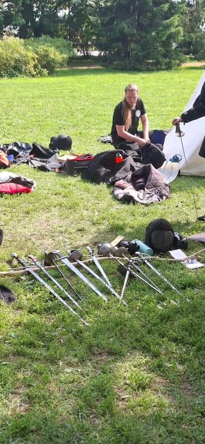 A group of people are engaged in a historical fencing practice session in a park, likely in Turku, Finland, given the provided context. In the foreground, several fencing foils and sabers are laid out on the grass, arranged in a line. These weapons appear to be part of the equipment for the practice. To the right, a person dressed in fencing attire, including a mask and gloves, is holding a foil. Behind the swords, there are various bags, mats, and protective gear scattered on the grassy ground, indicating preparation or aftermath of the activity. In the middle ground, a person with long blonde hair, wearing a black t-shirt, is seated on the grass, looking down at something in their hands, possibly adjusting equipment. The environment is a sunlit park with lush green grass and trees in the background, suggesting an outdoor recreational area.