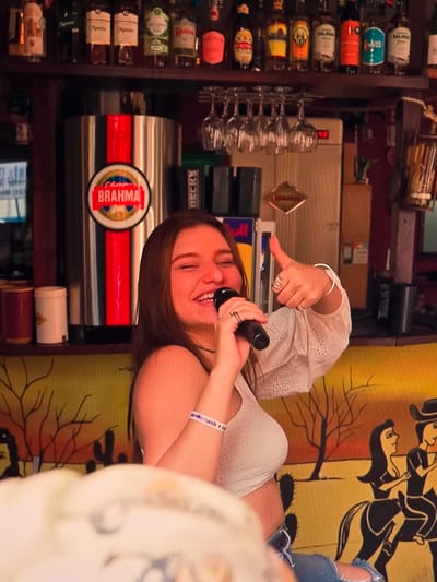 A young woman with long brown hair and a white crop top is holding a microphone and singing. She is smiling and giving a thumbs-up with her right hand, which is adorned with several silver rings and a bracelet. She is wearing blue jeans. Behind her, a Brahma beer dispenser is visible. The background shows a shelf with various bottles, wine glasses hanging, and a mural depicting a desert scene with cacti and people riding horses. The overall setting appears to be a bar or restaurant in Natal, Brazil, suggested by the presence of the Brahma brand, a popular Brazilian beer.