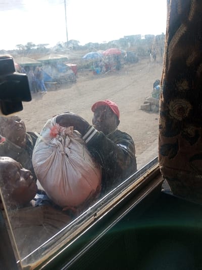 The image, taken from inside a vehicle looking out through a scratched window, depicts an active market scene in Kinanie ward, Kenya. In the immediate foreground, three adult males are engaged with a large, tightly-tied, light-colored sack at the vehicle's window opening. A man in a red baseball cap and dark jacket with white striped sleeves is central, using both hands to maneuver the sack, appearing to load or unload it. Another man to his left assists with the sack, while a third person's face is partially visible further left. The background reveals an expansive, dry, unpaved market area with multiple makeshift stalls. These stalls feature tarpaulins (blue, green) and large market umbrellas (red, blue). Several other individuals are visible in the mid-ground, scattered among the stalls, some seated. A utility pole stands in the distance, with further structures visible on a raised horizon under bright daylight conditions.