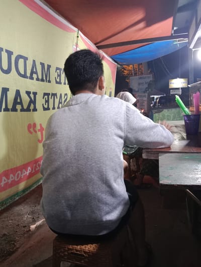 The image depicts a person seated with their back to the camera, facing a wooden table. The individual is wearing a grey long-sleeved shirt and dark shorts. They are positioned on a woven stool. The setting appears to be an outdoor food stall or warung, possibly in Sekopek Wetan, Indonesia. Behind the person, a large yellow banner with dark lettering is visible, with some text partially obscured. To the right of the table, a purple container holds green and pink plastic straws. The table surface is worn and appears to be damp in places. In the background, other people and signs are visible, suggesting a bustling environment. The lighting is dim, consistent with evening hours.