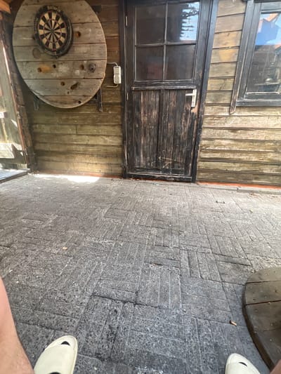 This image shows a partial view of a building exterior, likely located in Oud-Beijerland, Netherlands. The building has a wooden facade with a window and a wooden door or shutter. A round, wooden spool with a dartboard attached to it is mounted on the wall. The pavement is made of grey bricks. Part of a person, specifically their legs and feet clad in white slip-on shoes, are visible at the right side of the frame.