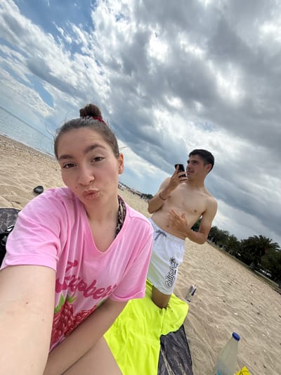 On a sandy beach in Νέα Φλογητά, Greece, a young woman in a pink t-shirt with a raspberry design is taking a selfie, her hair pulled back in a bun. Behind her, a young man in white shorts is looking at a phone. They are sitting on a beach blanket, and a water bottle and drink container are nearby. The sky is cloudy and the sea is visible in the background.
