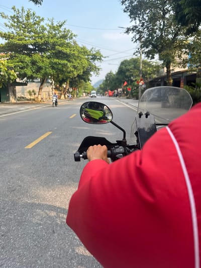 The image presents a first-person perspective from someone riding a motorcycle on a paved road during the daytime in Vị Xuyên Commune, Vietnam. The rider's right arm and torso, clad in a red long-sleeved garment with a white stripe, are prominent in the foreground. Their hand is seen gripping the motorcycle's handlebar and throttle.
The motorcycle's clear windshield is visible, and a smartphone is mounted centrally on the handlebars. The left rearview mirror reflects a person wearing a bright yellow or lime green jacket, likely another individual on a motorcycle or a passenger.
The road ahead features a solid yellow lane marking. Lush green trees line both sides of the road, creating a verdant tunnel effect. Beyond the trees, glimpses of buildings and utility poles with overhead wires can be seen. Small red flags, consistent with the design of the Vietnamese flag (red with a yellow star), are visible hanging among the trees in the distance, providing a strong geographical cue.
The bright sunlight indicates clear weather, casting shadows from the motorcycle and surrounding elements onto the road surface. The scene depicts an everyday activity of traveling by motorcycle through a tree-lined thoroughfare in a Vietnamese commune. There is no discernible text visible within the image.