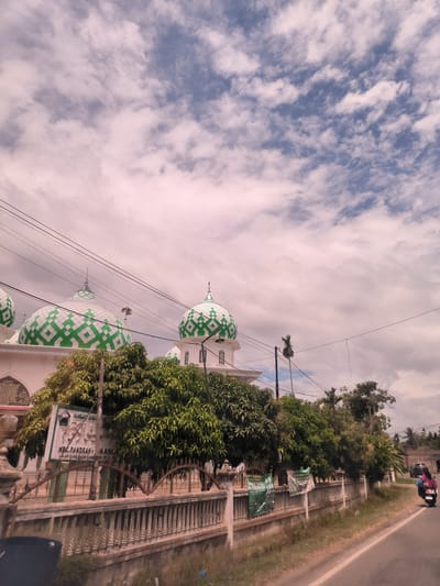 This image captures a roadside scene in Pandrahkandeh, Indonesia, featuring a mosque under a cloudy sky during the daytime. The most prominent subjects are the mosque's domes, which are distinctively patterned with green and white geometric squares. The mosque building itself is white, partially obscured by a line of dense green trees, likely mango trees, that separate the mosque compound from the road.
In the foreground, a concrete fence with decorative metal railings runs parallel to the asphalt road. Numerous power lines crisscross the upper portion of the frame. On the left, a sign provides specific location details, reading "MASJID BESAR..." (Grand Mosque), "KEC. PANDRAH - ID. BIREUEN" (Pandrah District, Bireuen Regency), and "JALAN BANDA ACEH MEDAN KM 170 PIDIE JAYA," confirming the setting as being along the Banda Aceh-Medan road. Further along the fence, green banners are visible, one of which clearly displays "SANTRI BARU" (New Students).
On the right side of the road, a person wearing a pink/purple top and blue pants is seen riding a white scooter away from the viewer, indicating typical daily activity. The sky, while mostly covered with white and grey clouds, shows glimpses of blue, suggesting mild weather conditions. The overall scene depicts a calm, routine moment in a semi-urban or rural Indonesian landscape.