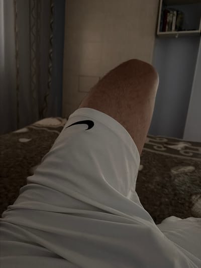 The image shows a close-up view of a person's leg, likely the thigh and part of the knee, wearing white athletic shorts with a black Nike logo. The leg is covered in light hair. The person appears to be lying down on a bed with a patterned blanket. In the background, a curtain is visible on the left, and a shelf with books is on the wall to the right. The scene is dimly lit, suggesting it might be indoors during the evening or in a room with limited natural light. The location is identified as Avdarma, Moldova.