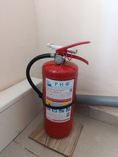 A red, cylindrical fire extinguisher stands upright on a light brown wooden mat on a tiled floor. The extinguisher has a black hose attached to its top, which loops around the body. The top of the extinguisher is silver with a red lever and handle. A white label is affixed to the body of the extinguisher, featuring Russian text and symbols, including a logo with stylized blue wings. The background consists of a plain, light-colored wall that appears to be slightly textured and a gray pipe running horizontally behind the extinguisher. The overall scene is indoors, in what appears to be a public or commercial space.