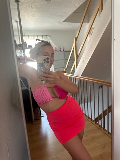 The image shows a young woman taking a mirror selfie in what appears to be a domestic setting, possibly a home in Heinebach, Germany. The woman is wearing a pink ruched bikini top and a bright orange-pink mini skirt. She is holding up her smartphone to capture the reflection. The background of the mirror reveals a staircase with wooden banisters and metal railings, and wooden flooring. The room appears to be well-lit, suggesting it is daytime. There are various objects visible in the background, including what looks like a shelf unit with items on it and some storage containers. The woman's expression is not clearly discernible as she is looking at her phone. There is no discernible text in the image, apart from what might be visible on the phone screen which is too blurry to read.