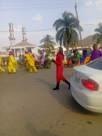 The image depicts a daytime street scene in Itakpe Junction, Nigeria, under a hazy sky with scattered clouds. The scene is observed from a slightly elevated perspective, looking across a paved road.

In the immediate foreground, the rear passenger side of a white sedan is visible, showing a red taillight and a green license plate partially obscured, displaying "SGB-883". A male figure, wearing a red long-sleeved shirt and matching red trousers with black stripes on the outer seams, and black socks, stands facing left, partially behind the white car, with his left foot slightly raised and his right foot on the asphalt. He has dark, dreadlock-like hair. His arms are bent at the elbows, with hands positioned in front of his torso, palms slightly open as if gesturing or speaking.

In the midground, to the left of the figure in red, a procession of individuals in vibrant yellow traditional attire, featuring elaborate head coverings often adorned with pink elements, moves from left to right across the road. At least four such individuals are clearly visible. Interspersed among them are several individuals on motorcycles, including one motorcycle carrying multiple passengers. One person on a motorcycle appears to be holding a green object resembling a decorated branch or standard. Further to the right, an orange motorized tricycle (keke napep) is visible, occupied by at least three individuals, with the driver in a green shirt and a passenger in a blue garment. To the right of the tricycle, another male figure in a purple garment stands, looking towards the camera.

In the background, on the far side of the road, a white mosque with two tall, slender minarets is prominently featured. The mosque has a light-colored roof and appears to be a multi-story structure. To the right of the mosque, several lush green palm trees are visible, with one large palm tree dominating the right side of the background. Other residential or commercial buildings with light-colored walls and roofs are also present. Overhead, power lines span across the frame.