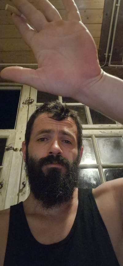 A man with a beard is looking at the camera, with his hand held up in front of his face. He is wearing a black tank top. Behind him are wooden panels and a window with multiple panes. The scene appears to be indoors, possibly in a rustic setting, with the background suggesting a location in Wattenwil, Switzerland, indicated by the wooden interior and style of the window, which might be characteristic of older Swiss architecture or a rural dwelling. The lighting is dim, with the hand obscuring a portion of the man's face.