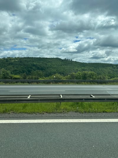 The image shows a view of a highway embankment with a dense green forest in the background. The sky is overcast with dramatic, dark clouds. On the highway, there are two lanes visible, separated by a white line, and a metal guardrail runs along the side. A narrow strip of green grass and a second guardrail are visible between the highway and the forest. A single white signpost is visible on the embankment. The scene is static and depicts a natural landscape adjacent to a transportation route. The image is taken from a low angle, looking slightly upwards towards the hill. The location is identified as Weischlitz, Germany.