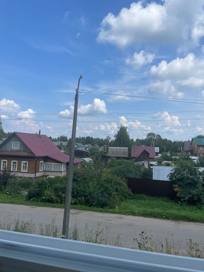 The image captures a view of a rural or suburban settlement on a clear, cloudy day. In the foreground, a gray metal barrier runs horizontally across the bottom of the frame. To the right, a tall, gray utility pole with wires stretches upwards. A paved road with some dry grass and weeds along its edge occupies the middle ground, leading towards a cluster of houses. The houses are of varying sizes and architectural styles, many featuring wooden construction and pitched roofs. One prominent house in the left half of the frame is a two-story wooden structure with a burgundy-colored roof, white window frames, and a porch area with decorative lattice work. Further back, a dense line of trees forms a backdrop to the settlement, with more scattered trees visible throughout. The sky above is a vibrant blue, punctuated by scattered white cumulus clouds. The overall impression is a peaceful, sunlit scene in a residential area. The location appears to be in Kotlas, Russia, based on visual cues consistent with the region's typical rural settlements.