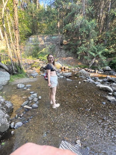 A full shot photograph, taken outdoors, captures a young woman standing in a shallow, rocky creek. She is positioned in the lower right quadrant of the image, facing left and looking towards the camera. She has long blonde hair and is wearing glasses, a black crop top, and light-colored shorts. A small backpack with red, white, and blue accents is visible on her back. Her feet are submerged in the clear water, and she is wearing light-colored sandals.
The creek bed is filled with a mixture of small pebbles and larger rocks of various sizes, some of which are partially submerged. The water is clear, allowing the creek bed to be visible. Sunlight reflects off the water's surface, creating bright spots.
The background features a natural, wooded environment. Tall evergreen trees with dense foliage dominate the upper portion of the image, with sunlight filtering through the branches. The ground slopes upwards behind the creek, covered in loose rocks and dirt, with some evidence of man-made structures, possibly retaining walls, visible on the embankment, which appear to be graffitied. Fallen logs and branches are scattered along the creek and on the banks. The overall setting suggests a natural area within a mountainous or forested region. The geographical coordinates indicate this scene is located in the electoral area of East Central Okanagan, British Columbia, Canada.