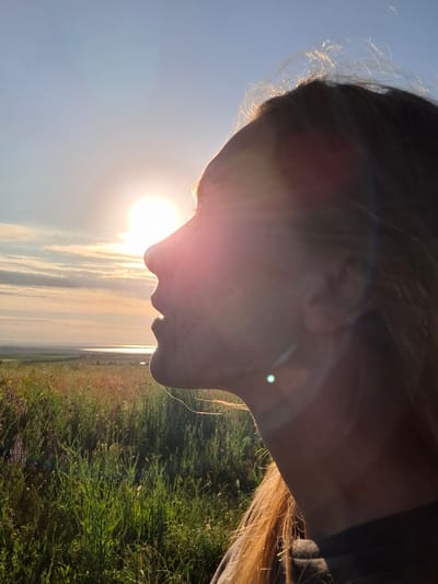 A profile view of a young Caucasian person's face is captured against a sunset in Akhtanizovskaya, Russia. The person is looking towards the left, their face illuminated by the setting sun which is positioned in the upper left quadrant of the image. The sun is bright and slightly diffused by clouds, casting a warm glow and creating lens flare effects. The person's hair, a light brown color, is blowing in the wind, with some strands catching the sunlight. The background consists of a vast, open field filled with green grass and small purple flowers, stretching towards a distant body of water and a hazy horizon. The overall mood is serene and contemplative, suggesting a moment of peace amidst nature.
