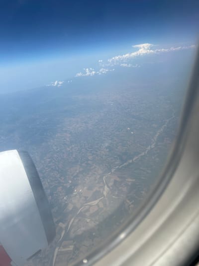 The image is an aerial view, likely taken from an airplane window, looking down at the landscape. The scene is dominated by a vast expanse of land divided into agricultural fields, suggesting a rural or semi-rural setting. A winding river snakes through the fields, indicating the presence of a significant watercourse. The land appears to be cultivated, with varying shades of brown and green indicating different crops or soil types. Some small structures, possibly houses or farm buildings, are scattered across the landscape, but they are too small to discern details.

In the upper left portion of the image, the distinct blue gradient of the sky is visible, transitioning into a lighter haze near the horizon. Wispy clouds are present, both along the edge of the atmosphere and scattered over the land. A portion of the airplane’s wing or fuselage is visible in the lower right corner of the image, appearing as a smooth, light-colored surface, further confirming the aerial perspective.

The overall impression is one of a bright, clear day, likely sometime in the late morning or early afternoon, given the quality of light and the sharpness of the shadows, though shadows are not prominent in this particular angle. The location is specified as Pavia, Italy, which is consistent with the appearance of a European landscape with agricultural patterns and a river. There is no visible text in the image. The mood conveyed is one of peaceful observation of the earth from above.