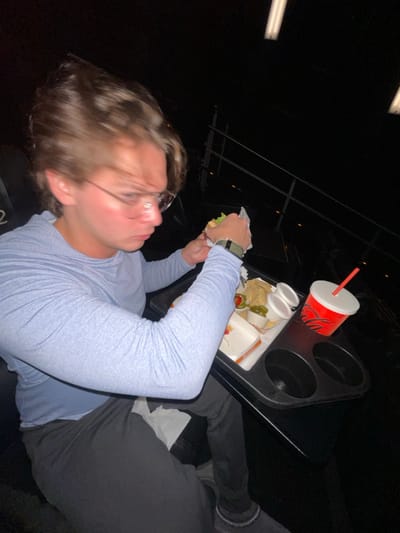 The image depicts one male individual seated within a dimly lit cinema environment in La Purísima, Mexico. The subject, a young adult with light brown hair and wearing clear-framed glasses, is dressed in a light blue long-sleeved hooded shirt and dark-colored pants. A dark smartwatch is visible on his left wrist. He is actively unwrapping or consuming a food item, consistent with a sandwich or burger, using both hands. His gaze is directed downwards towards the food.
A black tray is positioned in front of the male, featuring two circular cup holders and a flat serving area. On the tray, a red and white cup branded "Coca-Cola" with a red straw is placed in one cup holder. The serving area holds a white and orange paper container, likely a fast-food wrapper, and a portion of tortilla chips or nachos. Two small white plastic cups accompany the chips; one contains green sliced jalapeños, and the other appears to hold a white sauce or dip. A white paper napkin is partially visible on his lap and under his left hand.
The background is predominantly dark, indicating low light typical of a cinema. Visible environmental details include dark, padded cinema seats with armrests; one seat bears the alphanumeric designation "D2." A dark metal railing is visible further back, suggesting tiered seating. Overhead, two elongated, rectangular white fluorescent light fixtures are discernible against the dark ceiling.