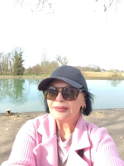 A middle-aged woman is depicted in a close-up, waist-up selfie portrait, positioned on what appears to be a canal bank in Souffelweyersheim, France. She wears a black baseball cap, tortoise-shell framed sunglasses, a light pink jacket with lapels, and a ribbed light pink long-sleeved top. A silver-colored necklace and gold-colored, padlock-shaped hoop earrings are visible. Her lips are painted a light pink. The woman directly faces the camera. Behind her, a calm body of water, likely a canal, reflects the bright sky and bare deciduous trees, appearing greenish-blue. On the canal bank to her left, a light-colored metal mooring bollard is present. The background features a line of bare deciduous trees, with a prominent conifer on the left. To the right, beyond the water, a light brown field and distant residential structures are visible under a bright, sunny sky. The overall lighting is strong natural light, indicating daytime.