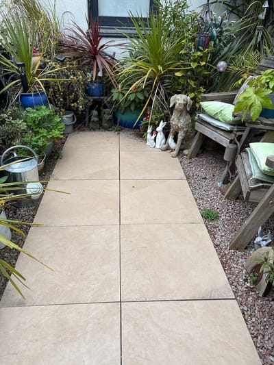 The image shows a garden path in Shrewsbury, United Kingdom, constructed of large, light-beige square paving slabs laid in a grid pattern. The path is flanked on one side by a densely planted garden area filled with various potted plants, including a cordyline plant with maroon leaves, various palms and other leafy plants, and a collection of garden ornaments. Among the ornaments are ceramic rabbits, a decorative dog statue, and several small elephant figures. A galvanized watering can sits near the edge of the path, partially obscured by foliage. Adjoining the paving slabs on the other side is a small area covered with reddish-brown gravel where a wooden chair is located with green cushions placed on it.