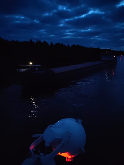 The image, taken at night, depicts a dark body of water with a long, dark barge illuminated by a single yellow light near the bow and a small red light on the stern. The opposite bank is lined with silhouetted trees. In the foreground is a white buoy with a red light. The sky is dark blue and cloudy. There is no evidence indicating that the image was taken in Uzhla, Russia.
