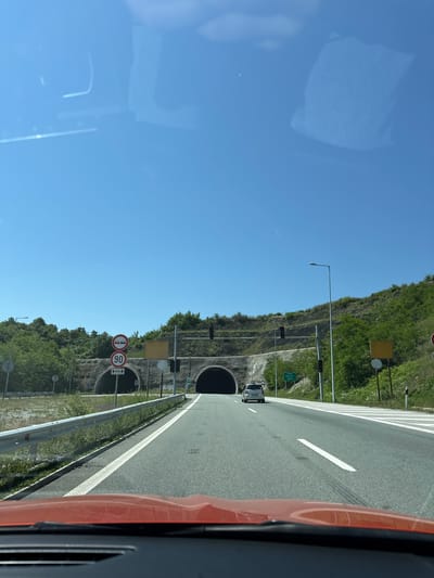The image presents a forward-facing view from inside a vehicle on a multi-lane asphalt highway, approaching a tunnel system under a clear blue sky. The scene is illuminated by bright sunlight.
In the foreground, the top of an orange/red vehicle's dashboard and windshield is visible. The road ahead is well-maintained, featuring white dashed and solid lane markings. A silver hatchback car is visible in the distance, driving in the same direction towards the central tunnel entrance.
To the left of the road, a metal guardrail runs alongside a grassy embankment. Further left, two circular traffic signs are stacked vertically: the upper sign depicts two red cars, indicating a "no overtaking" zone, and the lower sign displays "90" in a red circle, denoting a 90 km/h speed limit. Below the speed limit sign, an upward arrow with "400m" indicates the duration or distance for which the limit applies or the tunnel length. Another smaller, circular white sign with a diagonal black line, likely signifying the end of a previous restriction, is partially visible.
Two tunnel entrances are visible, carved into a tree-covered hillside. The main tunnel is centrally located ahead, with another tunnel entrance appearing to its left. Both entrances are reinforced with concrete or stone structures. Above the tunnels, traffic signals with dark housings are mounted, likely controlling access or indicating tunnel conditions.
On the right side of the road, a green rectangular sign with white Cyrillic text is visible. It reads "ТУНЕЛА БЛАТИНО" (TUNELA BLATINO) and "380M", explicitly identifying the location as the Blatino Tunnel and stating its length as 380 meters. This places the scene at the Blatino Tunnel in Blatino, Bulgaria. Adjacent to the sign is a street lamp. Empty square yellow signs are present on both sides of the road near the tunnel entrances. The hillsides surrounding the road and tunnels are densely covered with green trees and shrubs.