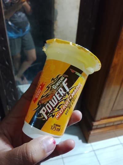A person is holding a yellow and orange plastic cup with a torn foil lid. The cup is branded with "POWER F" in large white letters, and the text "GINSENG" and "MIXFRUIT + MADU" are also visible. Below "POWER F", it reads "MENGANDUNG VIT B3 & B12". Further down, "ISI BERSIH 175 ml" indicates the net volume. The person's thumb has a small red mark on the nail. The background is blurred, showing what appears to be a reflection in a mirror of a person wearing a blue and grey shirt and grey shorts, and part of a wooden cabinet. The image appears to be taken indoors in a residential setting, possibly in Gunung Malang, Indonesia, given the product branding is in Indonesian.