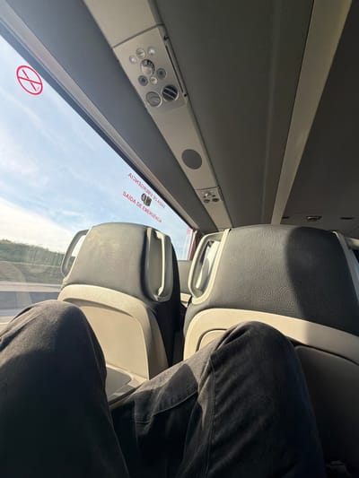 The image shows an interior shot of a bus or train, likely taken in Santarém, Portugal. The seating is upholstered, with a prominent view out of the window showing a landscape, possibly outside Santarém. A pair of legs clad in dark denim pants is visible occupying one of the seats. The window bears a "No Smoking" symbol and an emergency exit sign in Portuguese. The interior includes overhead controls and vents.