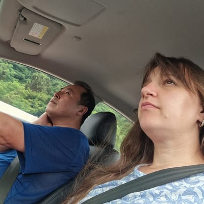 Two people, a man and a woman, are seated inside a car on a road in Jinbu-myeon, South Korea. The man, in the foreground and to the left, is wearing a blue striped shirt and is looking upwards and to his left, with his head resting on his neck. The woman, in the foreground and to the right, is wearing a blue and white patterned shirt and is looking upwards and to her right. Both are wearing seatbelts. The interior of the car is visible, including the ceiling, a sun visor with Korean text and an air bag warning sticker, and the car's headliner. Outside the car window, lush green trees are visible.
