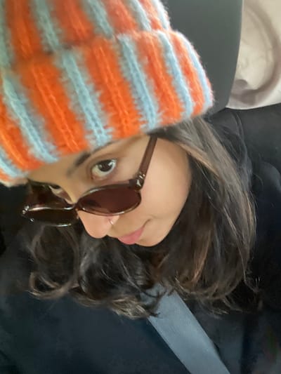 An individual is depicted seated within what appears to be the interior of a vehicle. The subject, presenting as female, is largely obscured by a close-up, high-angle shot. She is wearing a prominent orange and light blue striped knitted beanie. Dark brown sunglasses rest on her nose, partially obscuring her eyes, though her left eye (viewer's left) is visible above the frames, directed upwards and to the left. A small silver ring is present in her right nostril. Her lips are partially visible, showing a light pink coloration. She is wearing dark-colored attire, possibly a jacket or top, and a grey seatbelt is visible diagonally across her chest and shoulder. The background is indistinct, showing muted tones consistent with a vehicle's interior, including a dark element to the upper right, possibly a headrest or seat back. There are no visual cues within the image to confirm the location as the city of Elyashiv, Israel.