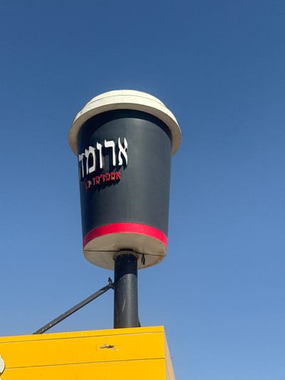 This media file shows a close-up, low-angle view of a cylindrical object against a clear blue sky. The object appears to be a part of a larger structure, possibly a sign or a decorative element. It is dark gray with white and red lettering. The white lettering is large and stylized, while the red lettering is smaller and appears to be in Hebrew.

The object has a wide, light-colored rim at one end and a red band closer to the middle. A black pipe extends from the object to the right, and a yellow structure is visible to the far right, suggesting an outdoor setting. The lighting indicates it is daytime, likely a sunny day given the clear sky.

The white lettering on the object is stylized and appears to spell out "XLIGHT". The red Hebrew lettering below it is harder to decipher due to the angle and resolution, but it may be a company name or a slogan. The overall impression is of a modern, possibly commercial, installation. The perspective suggests the object is mounted high up, and the viewer is looking upwards. The setting is likely Bnei Shimon Regional Council, Israel, based on the provided context.