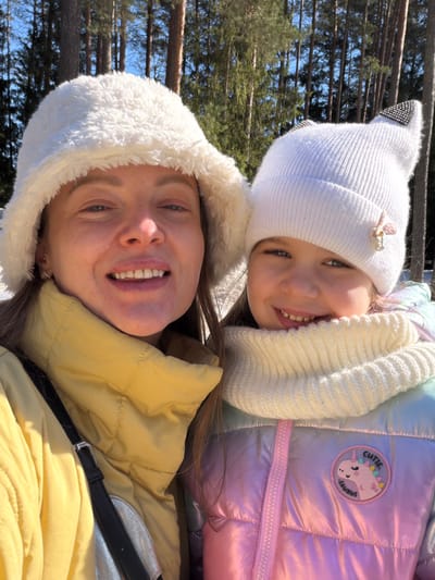 Two smiling females, an adult and a child, are depicted in a close-up, selfie-style outdoor portrait. The adult female, positioned on the left, wears a white, fluffy bucket hat and a light yellow puffer jacket. She has light brown hair partially visible, and small hoop earrings. The child, on the right, wears a white knit beanie with two pointed, black-studded "cat ears" and a light pink butterfly ornament. She also wears a white knit snood and an iridescent puffer jacket in shades of pink, purple, and blue, featuring a circular patch with a unicorn and the text "CUTIE. SAURUS". Both individuals are looking directly at the camera with broad smiles, revealing teeth. The background consists of numerous tall, slender tree trunks and patches of snow on the ground, indicating a forested, winter or early spring environment under a clear, bright blue sky. The scene is consistent with a natural, wooded area within Ракаўскі сельскі Савет, Belarus.
