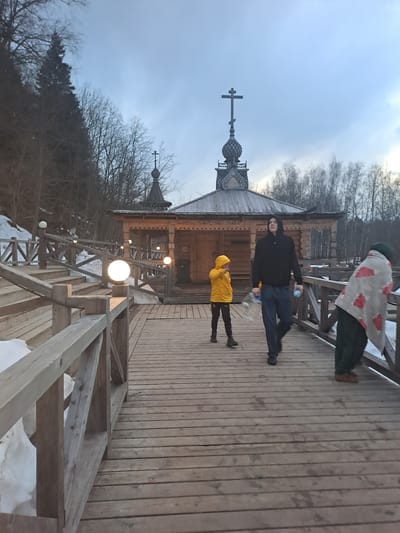 The image shows a wooden church in Vzglyadnevo, Russia, with a bridge leading to it. Three people are on the bridge: one in a green and red blanket, one in a black jacket and jeans, and one in a yellow hooded jacket. The bridge is wooden, and the church is constructed of logs, with a distinct onion dome topped with a cross. Several globe-shaped lights illuminate the bridge. Snow patches are visible near the base of the bridge supports. The sky is overcast.