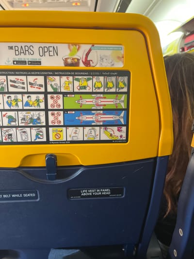 The image shows the back of an airplane seat. It has a yellow folding tray with safety instructions and information about Ryanair's bar service printed on it. Below the tray is the top of a navy-blue airplane seat. Text indicates the location of the life vest. The location, Somma Lombardo, Italy, is not apparent from the image.