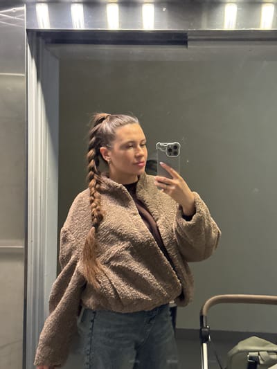 A female individual is depicted in a mirror selfie taken indoors in Gribovo, Russia. She stands facing a reflective surface, holding a gray smartphone with a distinctive light-colored, wavy-edged protective case in her right hand, angled towards her reflection. Her brown hair is styled in a high ponytail that transitions into a long braid extending over her left shoulder. She is wearing a light brown, textured, fuzzy bomber-style jacket over a darker brown top, paired with light blue denim jeans. Small hoop earrings are visible. Her head is slightly turned to her left, and her gaze is directed towards the phone screen. Overhead, three rectangular white lights are visible, illuminating the scene. A vertical metallic panel borders the left side of the reflective surface. In the lower right quadrant, the handle and a portion of a baby stroller or pram are visible, with a green or olive-drab bag resting nearby.