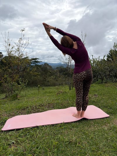 A woman is performing a yoga pose on a pink yoga mat outdoors. She is wearing a long-sleeved plum-colored top and leopard-print leggings. Her arms are raised above her head, palms together, and she is bending to her right, forming an arc with her body. The yoga mat is positioned on a grassy hill. In the background, there is lush green foliage, trees, and a hazy view of mountains in the distance under a cloudy sky. The location is identified as being in the city of Ban Khun Wang, Thailand.