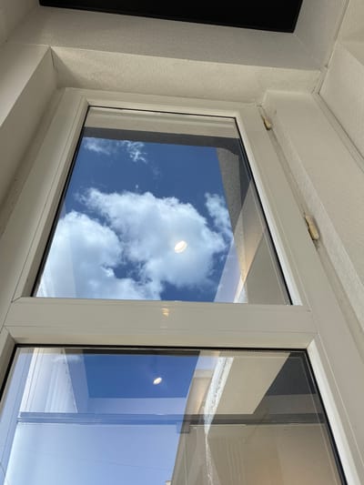 The image shows a close-up view of a white window frame, likely in the city of Khashuri, Georgia. The window reflects a bright blue sky with scattered white clouds. The reflection also captures a circular light fixture. The window itself appears to be divided into two panes, with the top pane showing the sky more prominently and the bottom pane reflecting the sky and some architectural elements, possibly buildings. The interior wall surrounding the window is painted a light color and has a textured wallpaper. A small, beige hinge is visible on the right side of the window frame, attached to the exterior wall. The scene is illuminated by natural daylight.