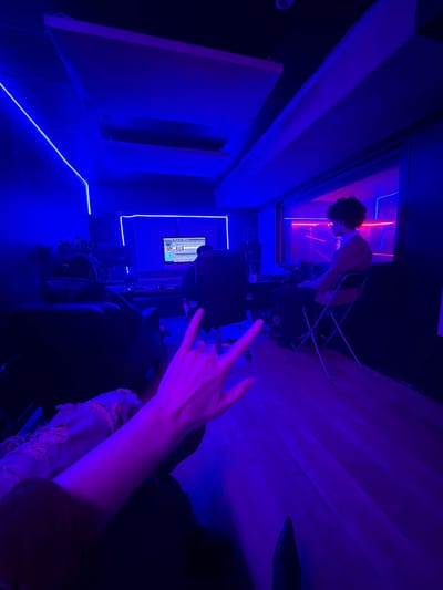 A dimly lit, upside-down room bathed in blue and purple hues. A person stands in a doorway with red neon lights above. In the foreground, a hand throws the "rock on" gesture. An adjacent room has blue neon accents and a small screen displaying graphics. Dark furniture and clothing obscure details, adding to the mysterious, edgy ambiance. The perspective creates a sense of disorientation and artistic expression.