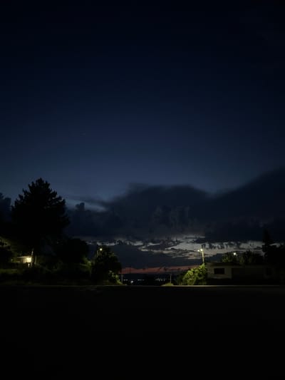 The image depicts a nighttime scene in Ivanski, Bulgaria, characterized by deep twilight and artificial illumination. The upper two-thirds of the frame are dominated by a dark indigo to navy blue sky, transitioning to lighter blues at the horizon. Prominent, dense, dark gray to black cumulus clouds traverse the mid-sky horizontally, with more fragmented, lighter gray clouds visible beneath them. A faint reddish-orange glow is present along the distant horizon. Scattered, minute white points, consistent with stars, are visible in the clearer sky sections.
The foreground, comprising the bottom third of the image, is a uniformly dark, unlit surface, likely an asphalt road or ground.
In the midground, on the left, a large, dark silhouette of a coniferous tree, likely a pine, stands prominently. Below this tree, a single-story building is partially visible, with a distinct warm yellow light source emanating from an interior opening or window. Further right, two street lights with bright, warm yellow illumination are visible; one illuminates a small, partially obscured building and adjacent foliage on the right side, while another is positioned further back on the left side, its light partially hidden.
The distant horizon features a subtle outline of undulating terrain or structures. On the far left of the horizon, a cluster of faint, twinkling yellow-orange lights indicates a distant settlement or city. A light-colored rectangular sign on a pole is barely discernible on the left side of the road, adjacent to the distant lights.
No human figures or explicit actions are discernible within the frame. The scene is illuminated by natural twilight and localized artificial street and interior lighting, resulting in deep shadows.