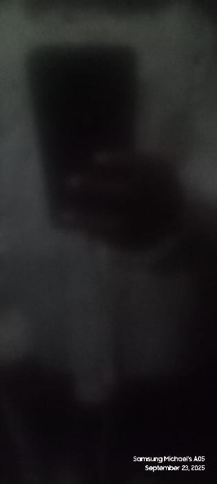The image, recorded by "Samsung Michael's A05" on September 23, 2025, is critically underexposed, blurry, and monochromatic in appearance, primarily composed of deep shadows and indistinct grey tones. In the upper portion, a large, dark, upright rectangular mass is discernible, possibly representing a piece of furniture or an architectural element. Below this, a heavily obscured, amorphous dark shape occupies the mid-ground, preventing identification of any specific subject, object, or person, or their actions. The overall environment appears to be dimly lit or completely dark, with no identifiable features such as walls, windows, or specific textures. The presence of people, explicit objects, or interactions cannot be determined from the visual data. The specific location within the city of Uweyoba, Nigeria, cannot be ascertained from the visual content provided in the image.